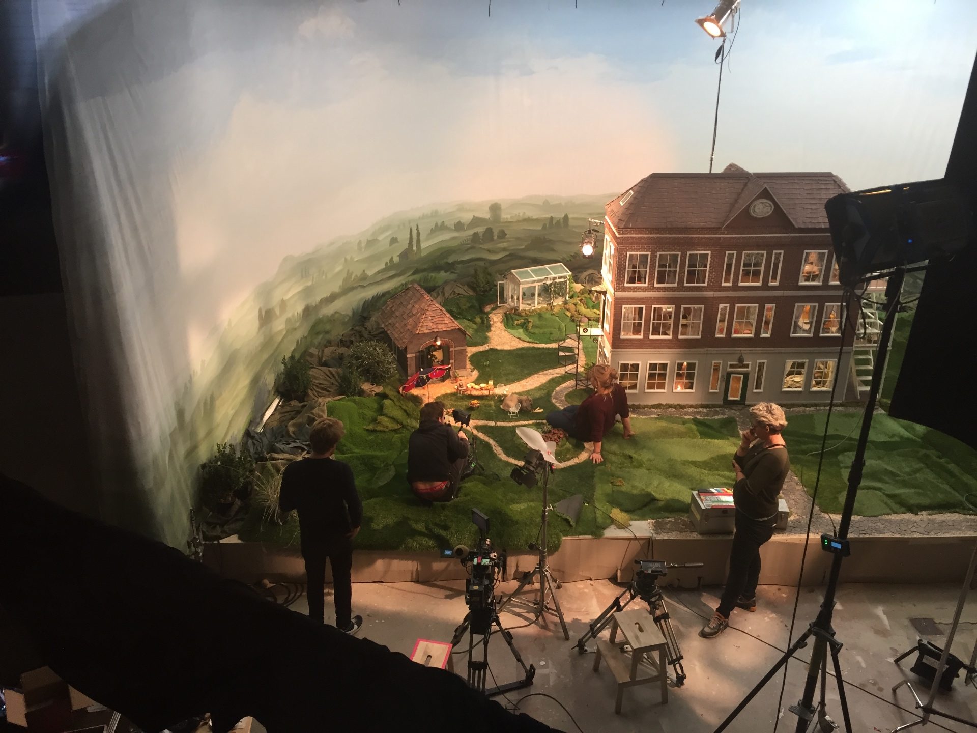 A wide shot of the studio: a miniature brick house and outbuilding on a sculpted hill in front of a painted landscape, with crew at human scale working around it